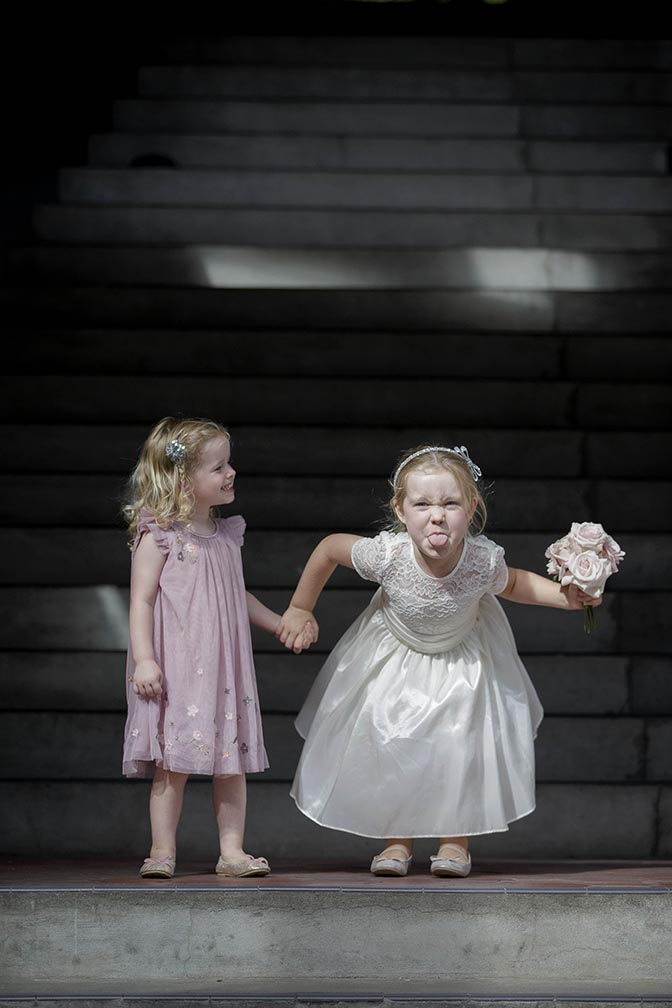 Little flower girl wearing white wedding style dress sticks her tounge out at the photographer whilst holding the hand of smiling flower girl wearing a pink dress. Auckland wedding photo by Chris Loufte at Cibo in the City. Capturing the moment.