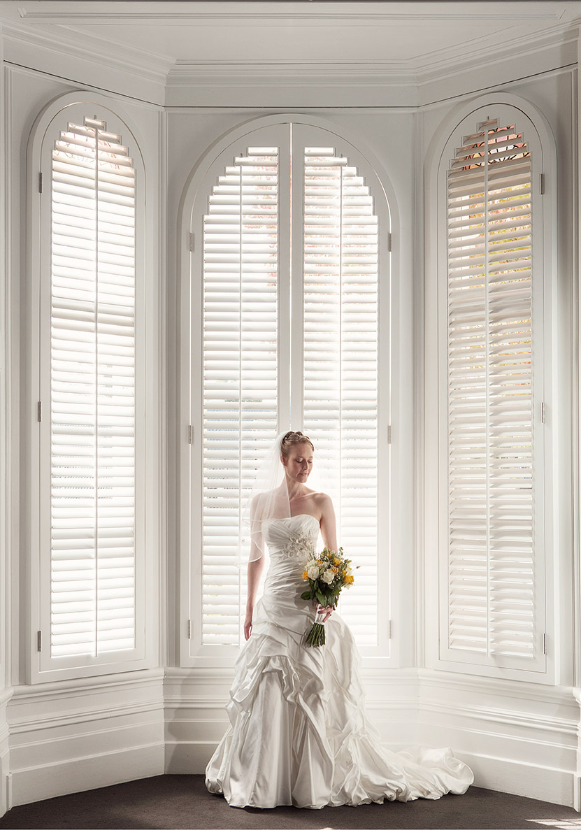 Wedding day bride wearing full length silk wedding gown holding a flower bouquet of yellow flowers standing in front of a double height ornate window with plantation shutters at Diocesan School Auckland.