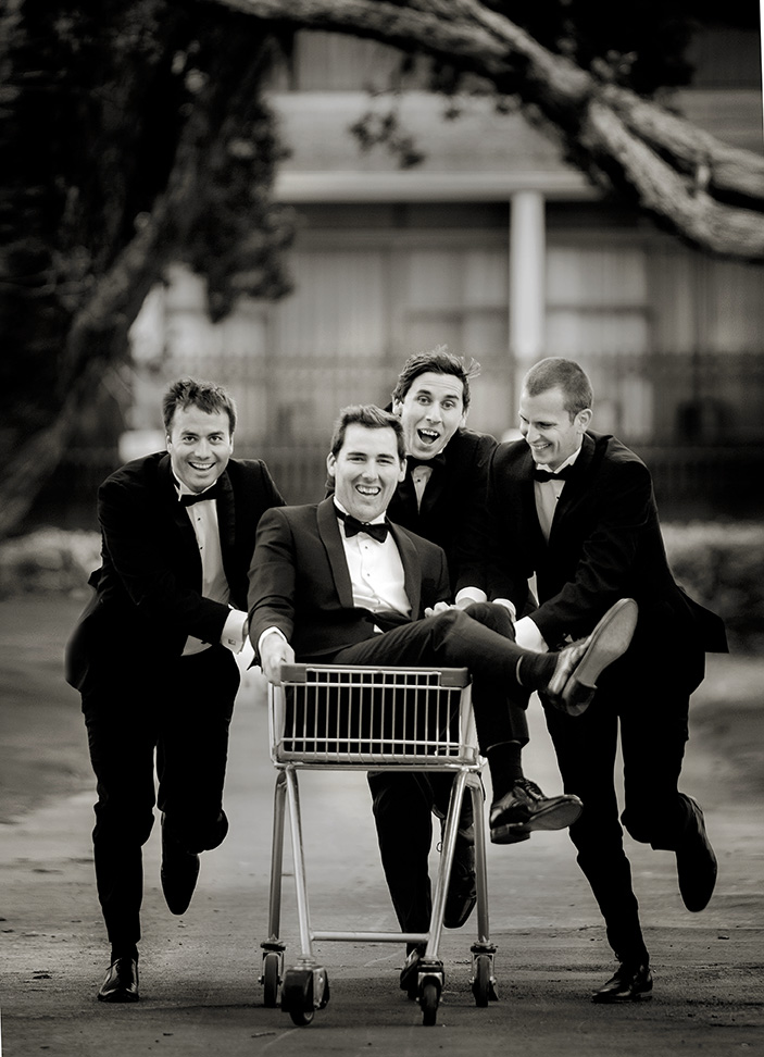 Three groomsmen dressed in matching black tuxedos pushing the groom in a shopping trolly along a road in Auckland as they get him to the church on time for his wedding at the Roman Catholic Cathedral of St PAtrick & St .joseph in Auckland. capturing the moment by Auckland Wedding photographer Chris loufte.