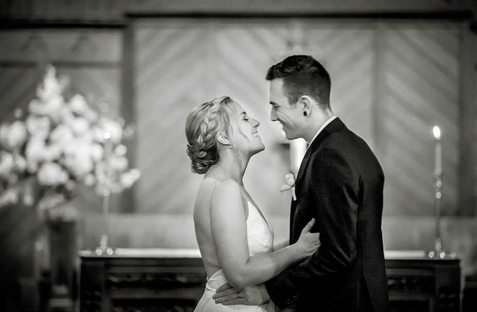 Bride wearing strapless white wedding dress and her new husband wearing a black suit look into each others eyes and smile in front of the alter at Saint Aidan's Church in Remuera in Auckland during their wedding ceremony. Capturing the moment by Auckland Wedding photographer Chris Loufte