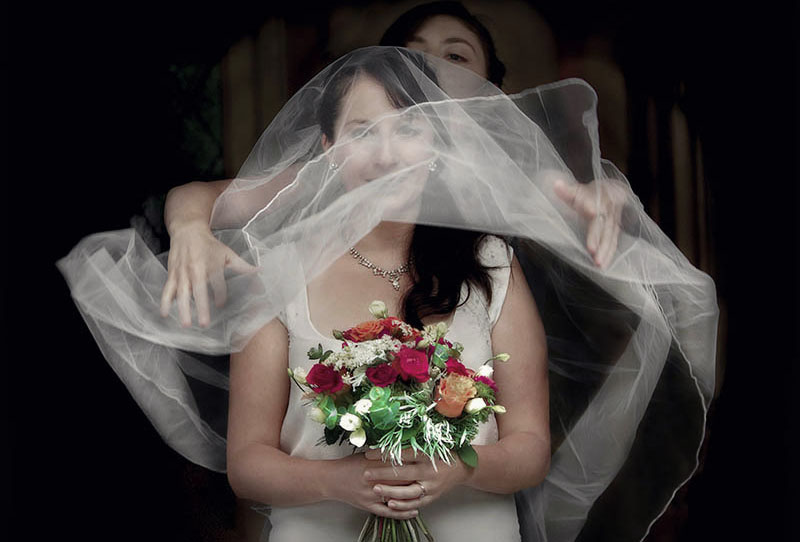 wedding day bride holding a brightly coloured flower bouquet has her veil thrown over her head in preparation for her ceremony.