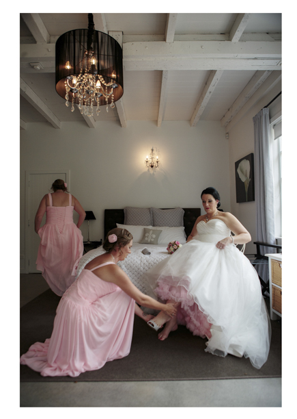 Bridesmaid in dusty pink pleated dress helps bride in full white dress to put her wedding shoes on.