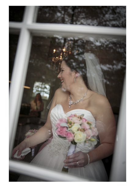 Bride wearing her wedding dress holding her floral bouquet photographed through window frame