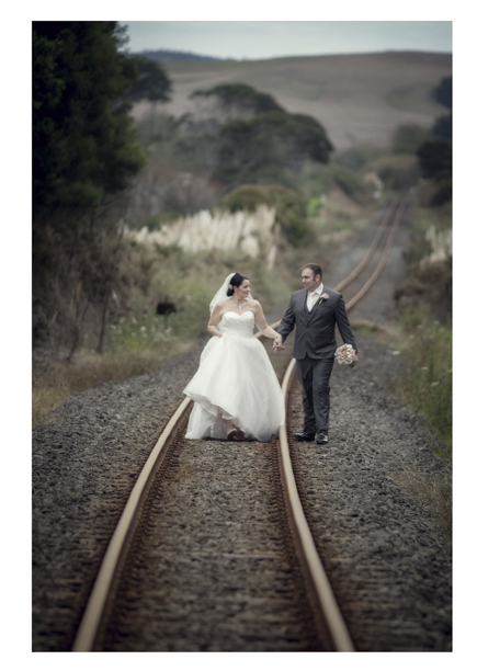 Bride in wedding dress walks with groom along romantic disused railway line in Kumeu.