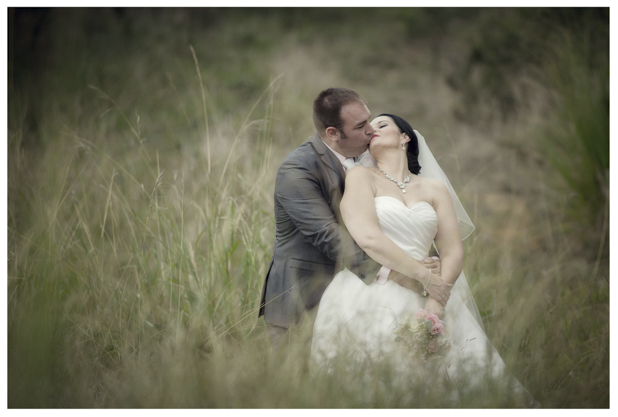 Bride in wedding dress embraces groom in long grass by a romantic disused railway line in Kumeu.
