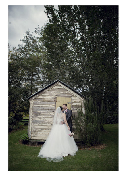 Bride in wedding dress embraces groom pose for a photo by rustic shed in Kumeu.