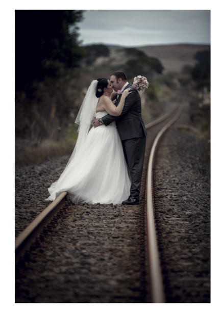 Bride in wedding dress embraces groom along romantic disused railway line in Kumeu.