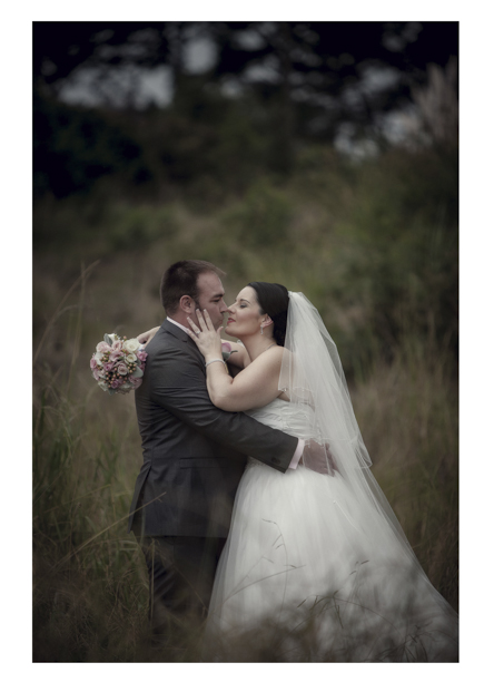 Bride in wedding dress embraces groom in long grass by a romantic disused railway line in Kumeu.