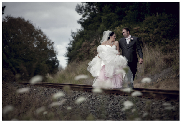 Bride in wedding dress walks with groom along romantic disused railway line in Kumeu.