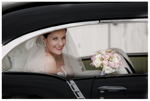 Bride holding flower bouquet sitting in the back of classic wedding car .