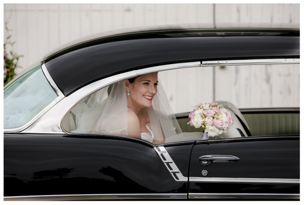 Bride holding flower bouquet sitting in the back of classic wedding car .
