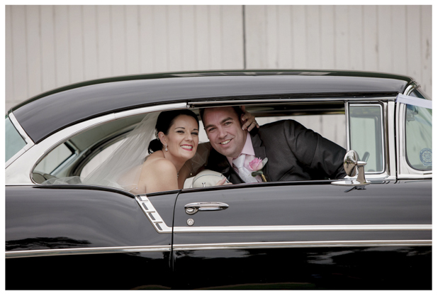 Bride holding flower bouquet and groom sitting in the back of classic wedding car .