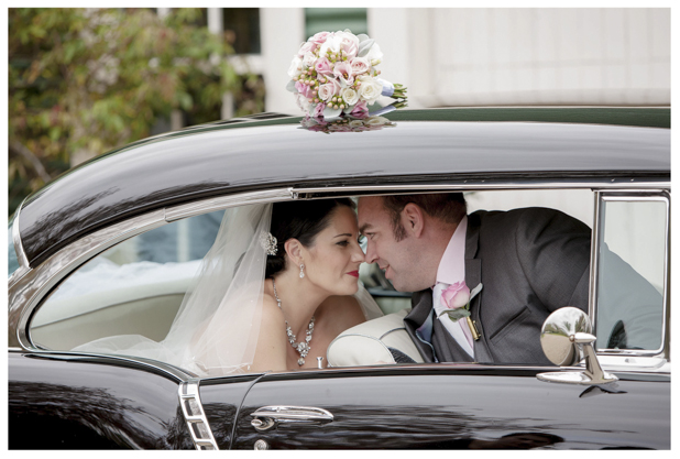 Bride holding flower bouquet and groom sitting in the back of classic wedding car .