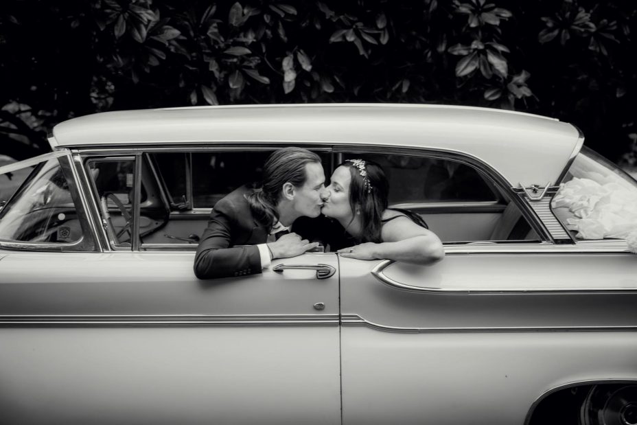 Bride and groom kiss in their classic American wedding car. Black and white photo