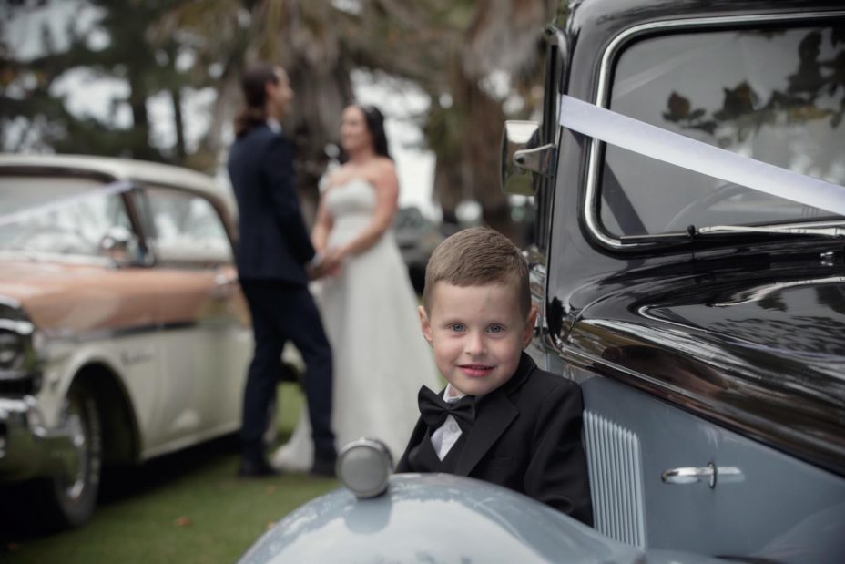 Cute pageboy leans on teh fender of classic wedding car