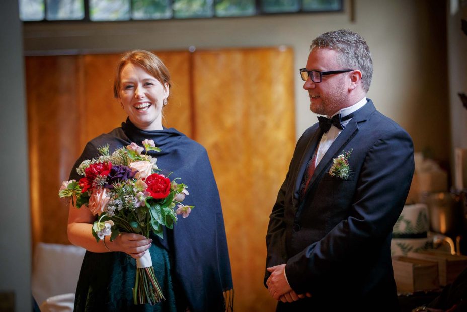 Bridesmaid holding flower bouquet laughs with a groomsman in formal black suit.