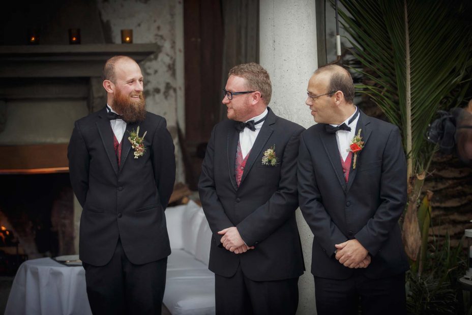 The groom and groomsmen wearing formal black suits and bow ties smile and chat together in front of the fireplace as they await the bride.