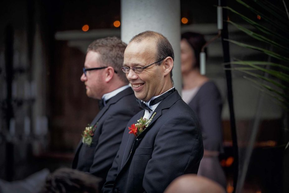 Groomsman wearing formal black bow tie and buttonhole flower smiles broadly at wedding guests.