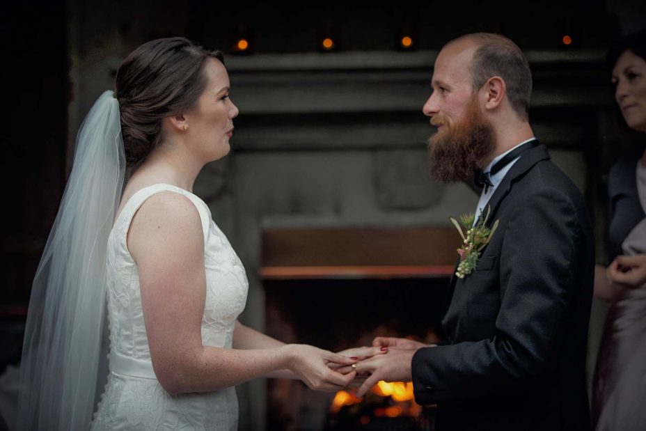 Bride pushes gold wedding ring into her groom's finger during their wedding ceremony in front of the fireplace at Mantells in Mount Eden.