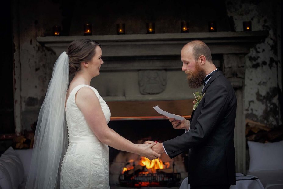 Smiling, happy bride and groom hold hands whilst groom reads his wedding vows in front of the fireplace at Mantells in Mount Eden.