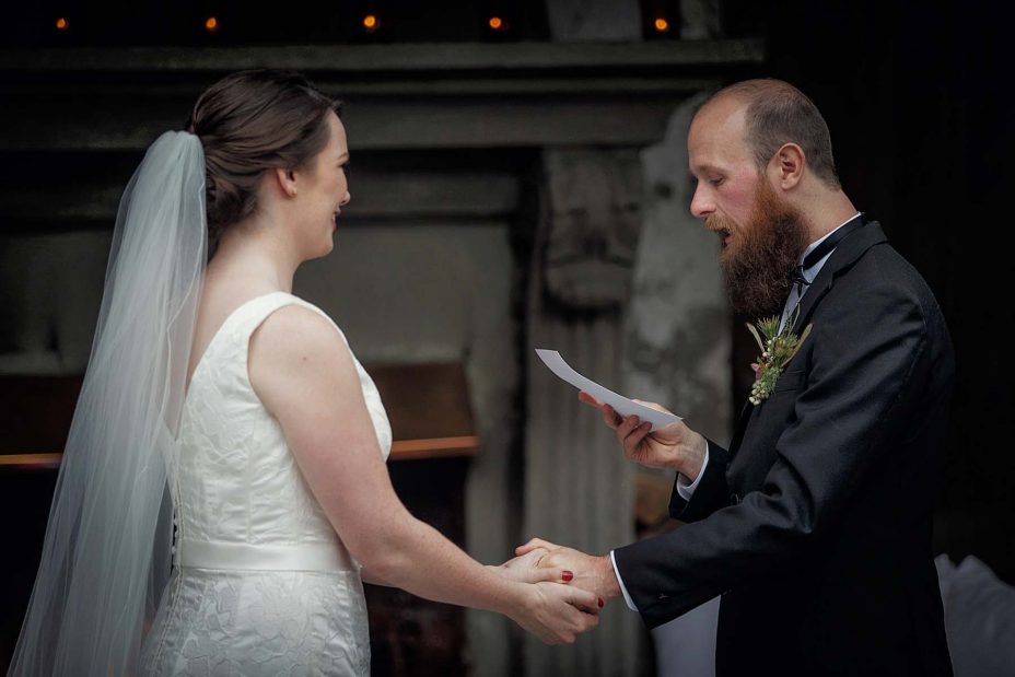 Smiling, happy bride and groom hold hands whilst groom reads his wedding vows in front of the fireplace at Mantells in Mount Eden.