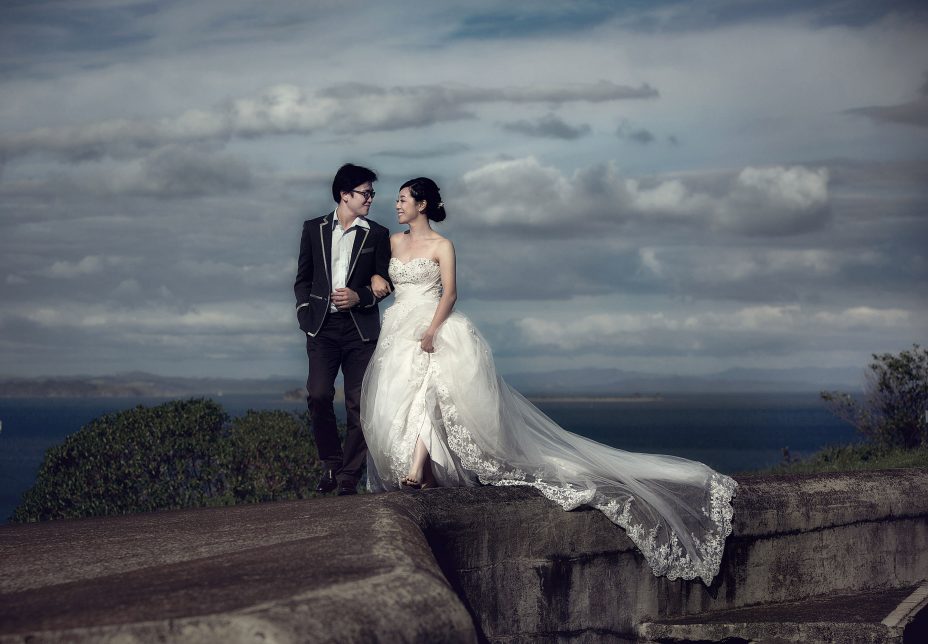 Chinese newlyweds walk arm in arm at North Head in Auckland. in the dramatic afternoon light. The bride wearing a full lenght white wedding dress with long gausey train trailing behind her. The couple share a special moment together captured by Auckland Wedding photographer Chris Loufte.