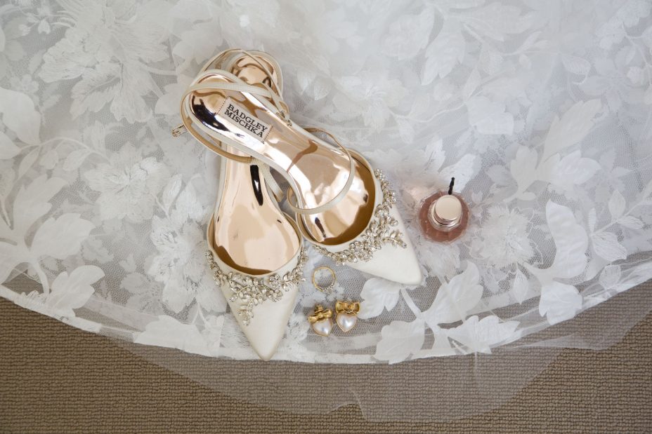 Wedding shoes with gold inner and floral diamante decoration on the toes sitting on he hem of a flower pattern white lace wedding dress alongside a diamond engagement ring and a pair of heart shaped pearl and gold earrings for the bride to wear to her wedding ceremony and reception at Fort Takapuna and the Officers Mess in Auckland.