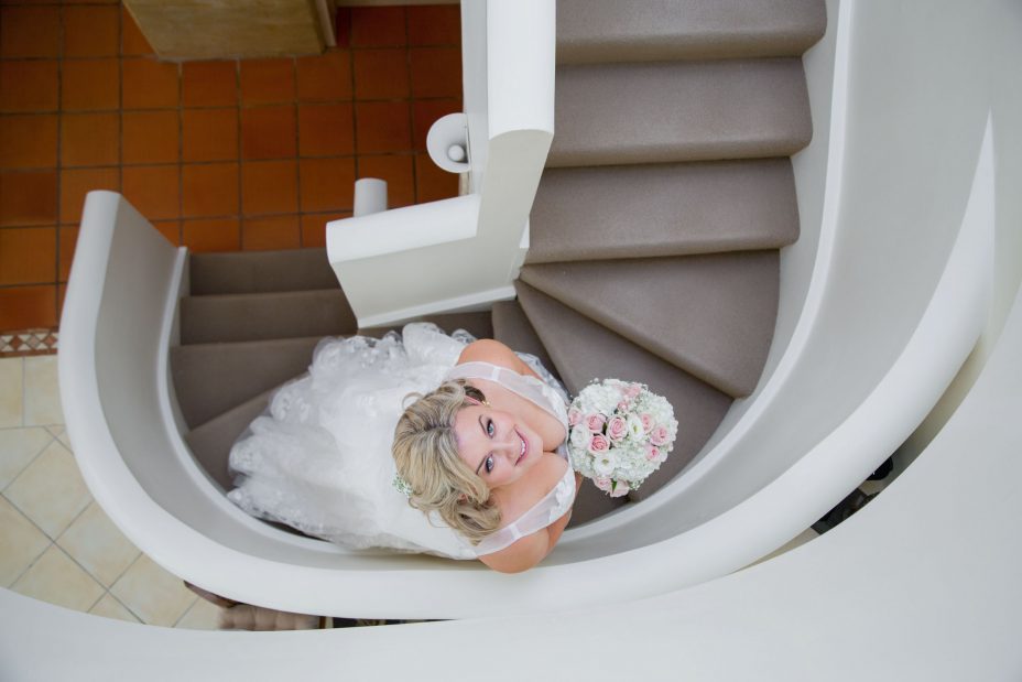 Bride in white flower pattern wedding dress holding a flower bouquet of pink and white roses looking up and smiling on an elegant curved staircase before her wedding at Fort Takapuna and the Officers Mess in Auckland.