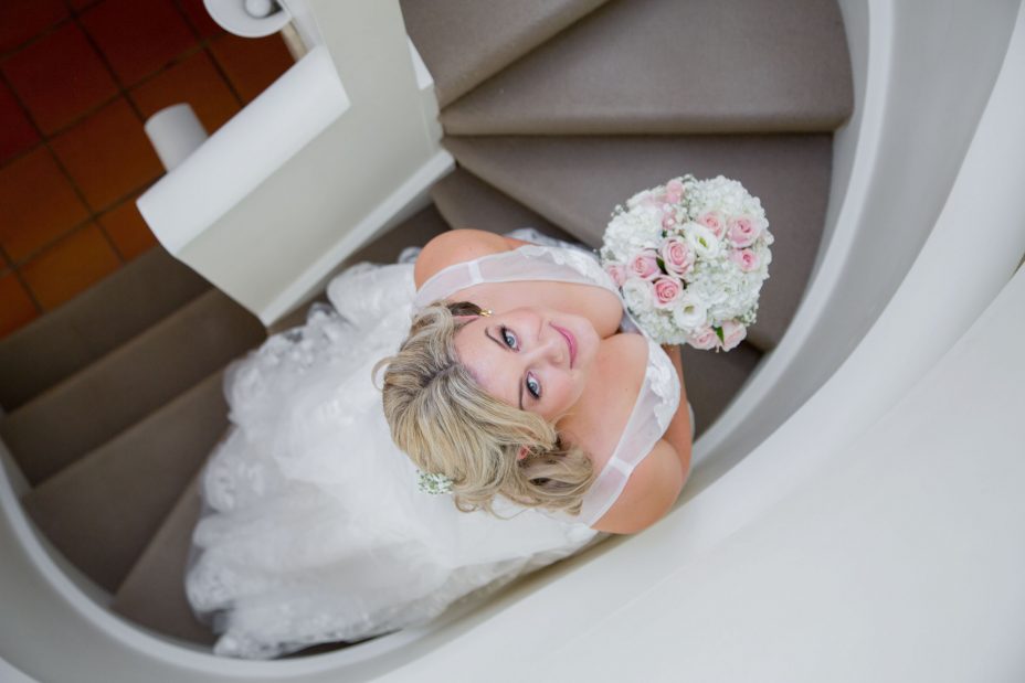 Bride in white flower pattern wedding dress holding a flower bouquet of pink and white roses looking up and smiling on an elegant curved staircase before her wedding at Fort Takapuna and the Officers Mess in Auckland.
