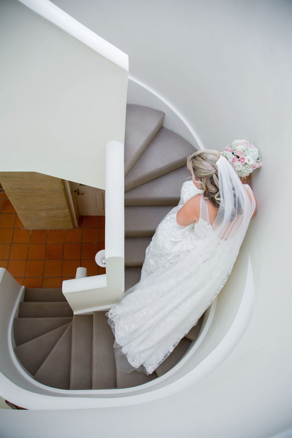 Bride in white flower pattern wedding dress holding a flower bouquet of pink and white roses looking up and smiling on an elegant curved staircase before her wedding at Fort Takapuna and the Officers Mess in Auckland.