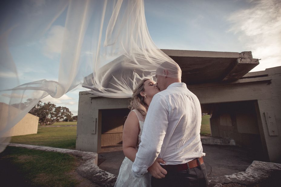 Sunset wedding photo of bride in white wedding dress with her newlywed husband kissing with her veil blowing around them at Fort Takapuna and the Officers Mess on a sunny evening in Auckland.