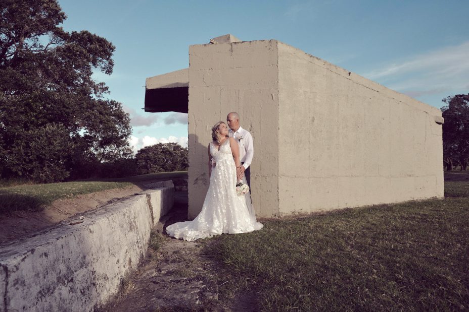 Bride wearing a full style white wedding dress holding her flower bouquet poses cuddled in with her new husband, for a creative wedding photo in front of a unique concrete structure at Fort Takapuna and the Officers Mess in Auckland