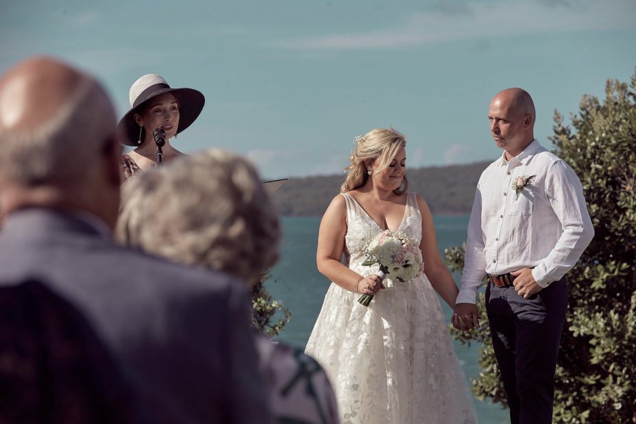 Wedding ceremony on a sunny day with a view of the sea and rangitoto Island. Guests watch as the celebrant reads to the bride wearing a full style white wedding dress and groom wearing white linen shirt and dark blue chino pants hold hands during their ceremony at Fort Takapuna and the Officers Mess on a sunny summer day in Auckland.