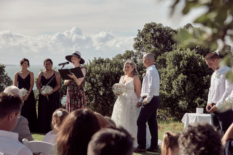 Wedding ceremony on a sunny day with a view of the sea and rangitoto Island. Guests watch as the celebrant reads to the bride wearing a full style white wedding dress and groom wearing white linen shirt and dark blue chino pants hold hands during their ceremony at Fort Takapuna and the Officers Mess on a sunny summer day in Auckland.