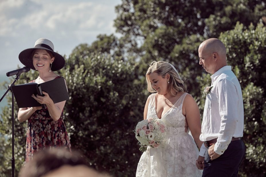 Wedding ceremony on a sunny day with a view of the sea and rangitoto Island. Guests watch as the celebrant reads to the bride wearing a full style white wedding dress and groom wearing white linen shirt and dark blue chino pants hold hands during their ceremony at Fort Takapuna and the Officers Mess on a sunny summer day in Auckland.