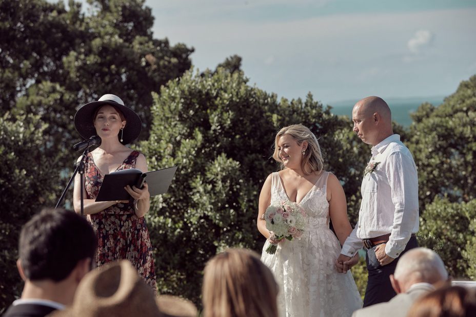 Wedding ceremony on a sunny day with a view of the sea and rangitoto Island. Guests watch as the celebrant reads to the bride wearing a full style white wedding dress and groom wearing white linen shirt and dark blue chino pants hold hands during their ceremony at Fort Takapuna and the Officers Mess on a sunny summer day in Auckland.