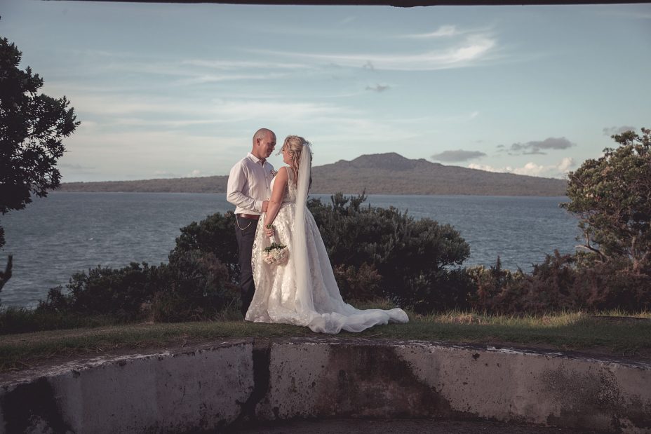A sunny day wedding with a view over the sea to Rangitoto Island in the background. Bride wearing a full style white wedding dress with long stands with her new husband on a unique concrete structure for a creative wedding photo at Fort Takapuna and the Officers Mess in Auckland
