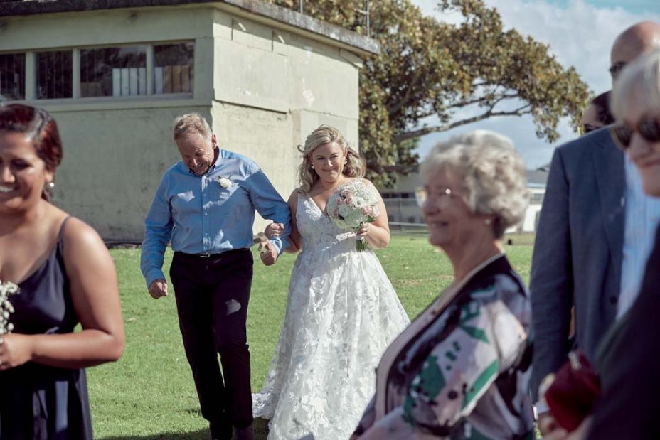 A bride wearing a full style white wedding dress walks arm in arm with her father down an isle of guests on a sunny day as she arrives at her wedding ceremony at Fort Takapuna and the Officers Mess in Auckland.