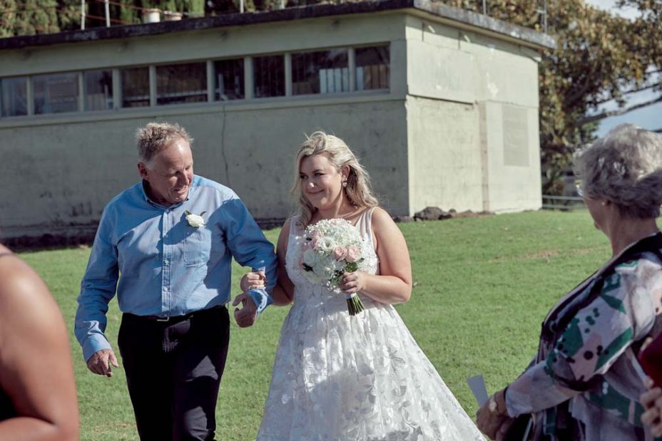A smiling bride wearing a full style wedding dress holding a pink and white rose flower bouquet walks arm in arm with her father down an isle of guests on a sunny day as she arrives at her wedding ceremony at Fort Takapuna and the Officers Mess in Auckland.