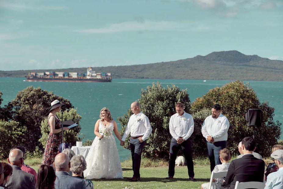 Wedding ceremony on a sunny day with a view of the sea and rangitoto Island. Guests watch as the celebrant reads to the bride wearing a full style white wedding dress and groom wearing white linen shirt and dark blue chino pants hold hands during their ceremony at Fort Takapuna and the Officers Mess on a sunny summer day in Auckland.
