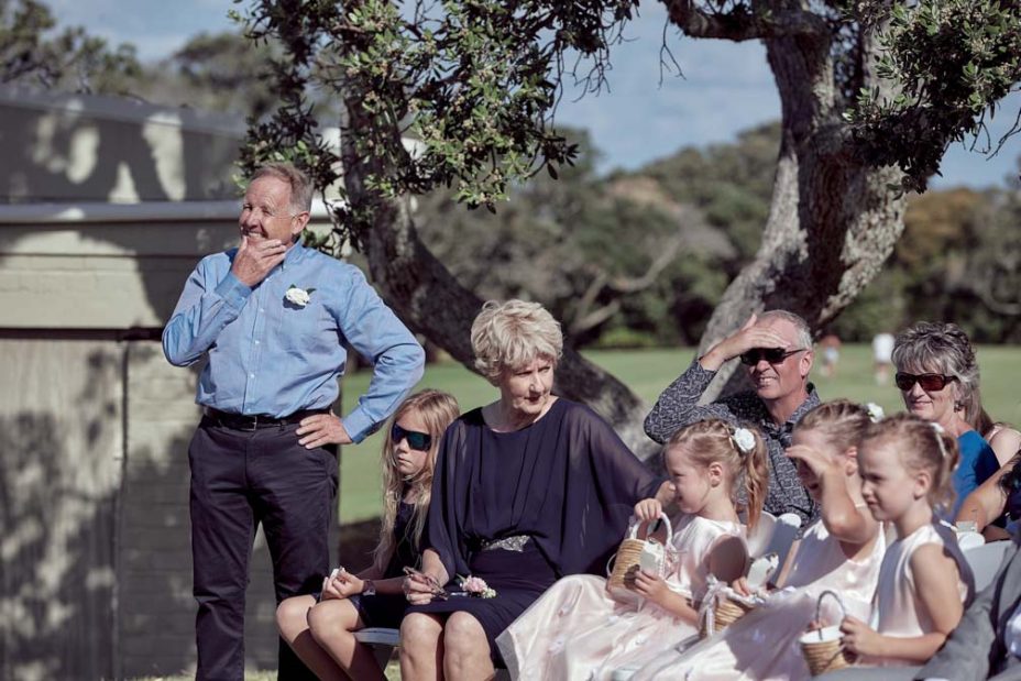 Father of the bride smiles with joy as he watches as the celebrant reads to the bride wearing a full style white wedding dress and groom wearing white linen shirt and dark blue chino pants making their wedding vows during their ceremony at Fort Takapuna and the Officers Mess on a sunny summer day in Auckland