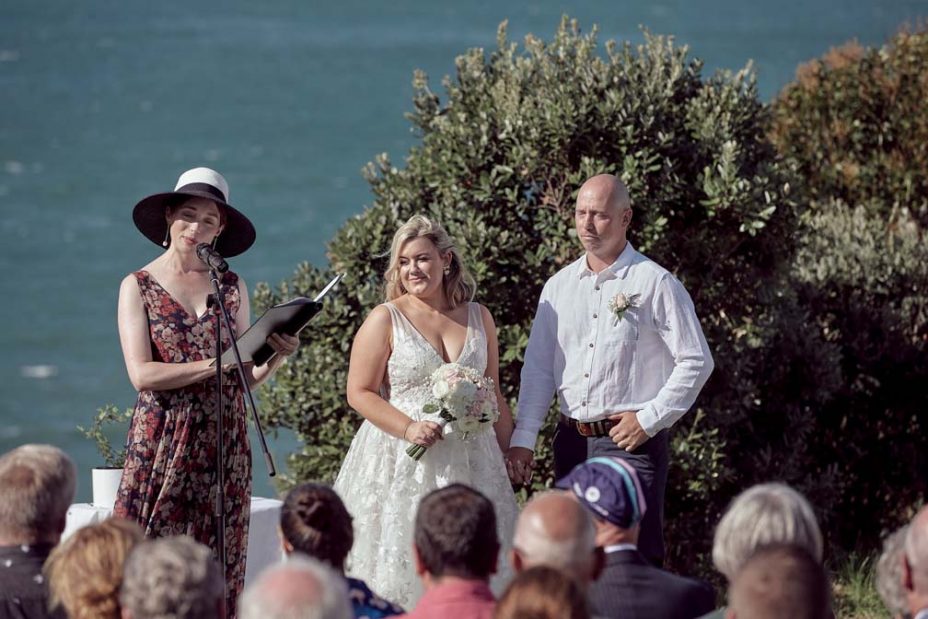 Wedding ceremony on a sunny day with a view of the sea and rangitoto Island. Guests watch as the celebrant reads to the bride wearing a full style white wedding dress and groom wearing white linen shirt and dark blue chino pants hold hands during their ceremony at Fort Takapuna and the Officers Mess on a sunny summer day in Auckland.
