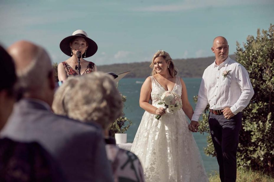 Wedding ceremony on a sunny day with a view of the sea and rangitoto Island. Guests watch as the celebrant reads to the bride wearing a full style white wedding dress and groom wearing white linen shirt and dark blue chino pants hold hands during their ceremony at Fort Takapuna and the Officers Mess on a sunny summer day in Auckland.