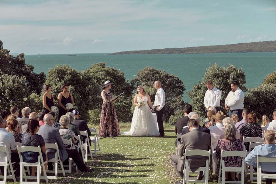 Wedding ceremony on a sunny day with a view of the sea and rangitoto Island. Guests watch as the celebrant reads to the bride wearing a full style white wedding dress and groom wearing white linen shirt and dark blue chino pants hold hands during their ceremony at Fort Takapuna and the Officers Mess on a sunny summer day in Auckland.
