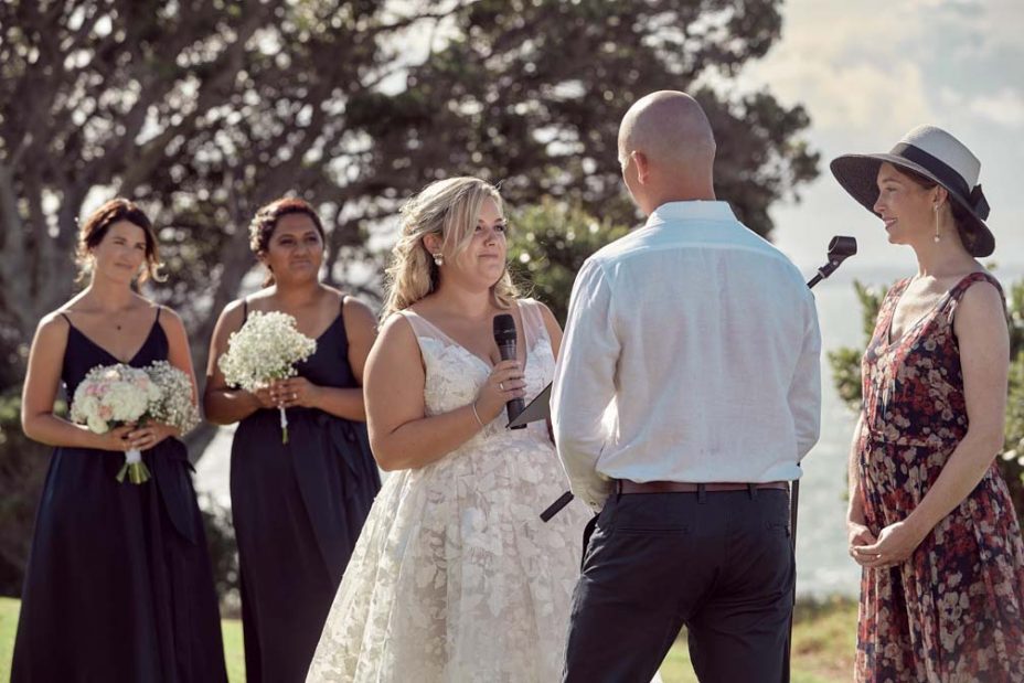 Wedding ceremony on a sunny day with a view of the sea and rangitoto Island. Guests watch as the celebrant reads to the bride wearing a full style white wedding dress and groom wearing white linen shirt and dark blue chino pants hold hands during their ceremony at Fort Takapuna and the Officers Mess on a sunny summer day in Auckland.