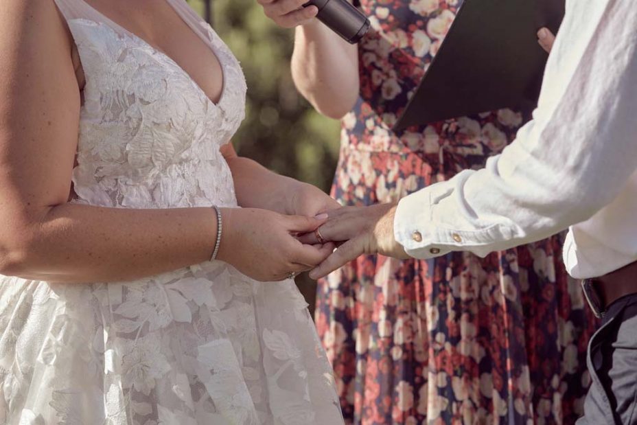 Close up photo of bride wearing a full style white wedding dress and groom wearing white linen shirt and dark blue chino pants placing a wedding ring on her new husband's finger during their wedding ceremony at Fort Takapuna and the Officers Mess in Auckland