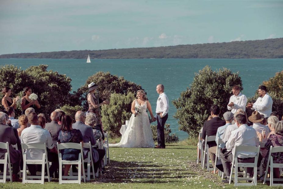 Wedding ceremony on a sunny day with a view of the sea and rangitoto Island. Guests watch as the celebrant reads to the bride wearing a full style white wedding dress and groom wearing white linen shirt and dark blue chino pants hold hands during their ceremony at Fort Takapuna and the Officers Mess on a sunny summer day in Auckland.