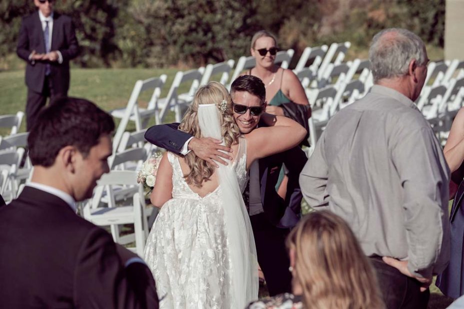 Bride hugs a wedding guest in amongst folding white chairs after her wedding ceremony at Fort Takapuna on a sunny summer day in Auckland.