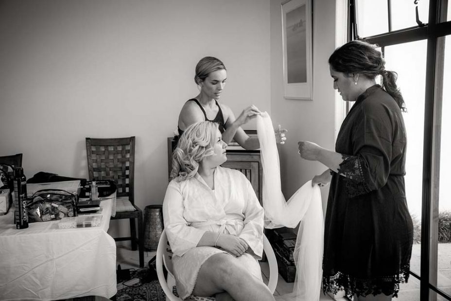 Black and white photo of a wedding day bride with a bridesmaid and a stylist as they stretch out her wedding veil to work out how to attache it to the bride's hair style. The bride sits in her pink robe as she prepares for her wedding ceremony and reception at Fort Takapuna and the Officers Mess in Auckland.