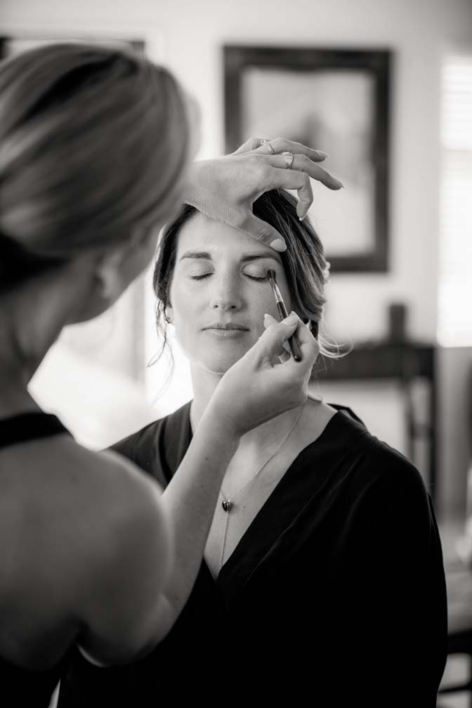 Black and white photo of a wedding day bridesmaid having her eye shadow applied with a soft brush during her makeup she gets ready for the wedding ceremony and reception at Fort Takapuna and the Officers Mess in Auckland.